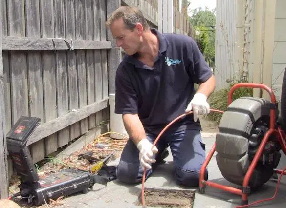 A plumber feeding a drain camera cable into a drain beside a fence